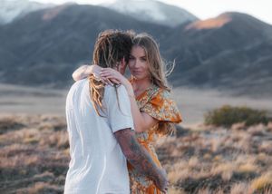 Couple hugging in a mountain field, man with dreadlocks and tattoos, woman in orange floral dress.