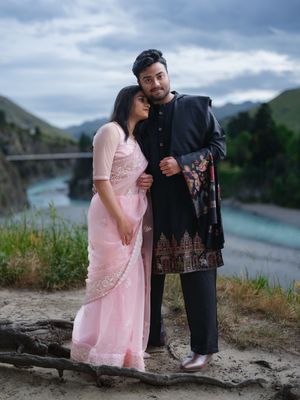 Couple in traditional South Asian attire posing by a river with mountains in the background.