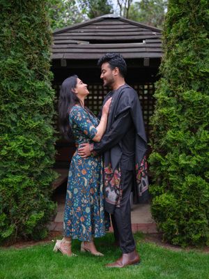A couple in traditional South Asian attire embraces lovingly in a lush garden with tall hedges and a wooden gazebo.