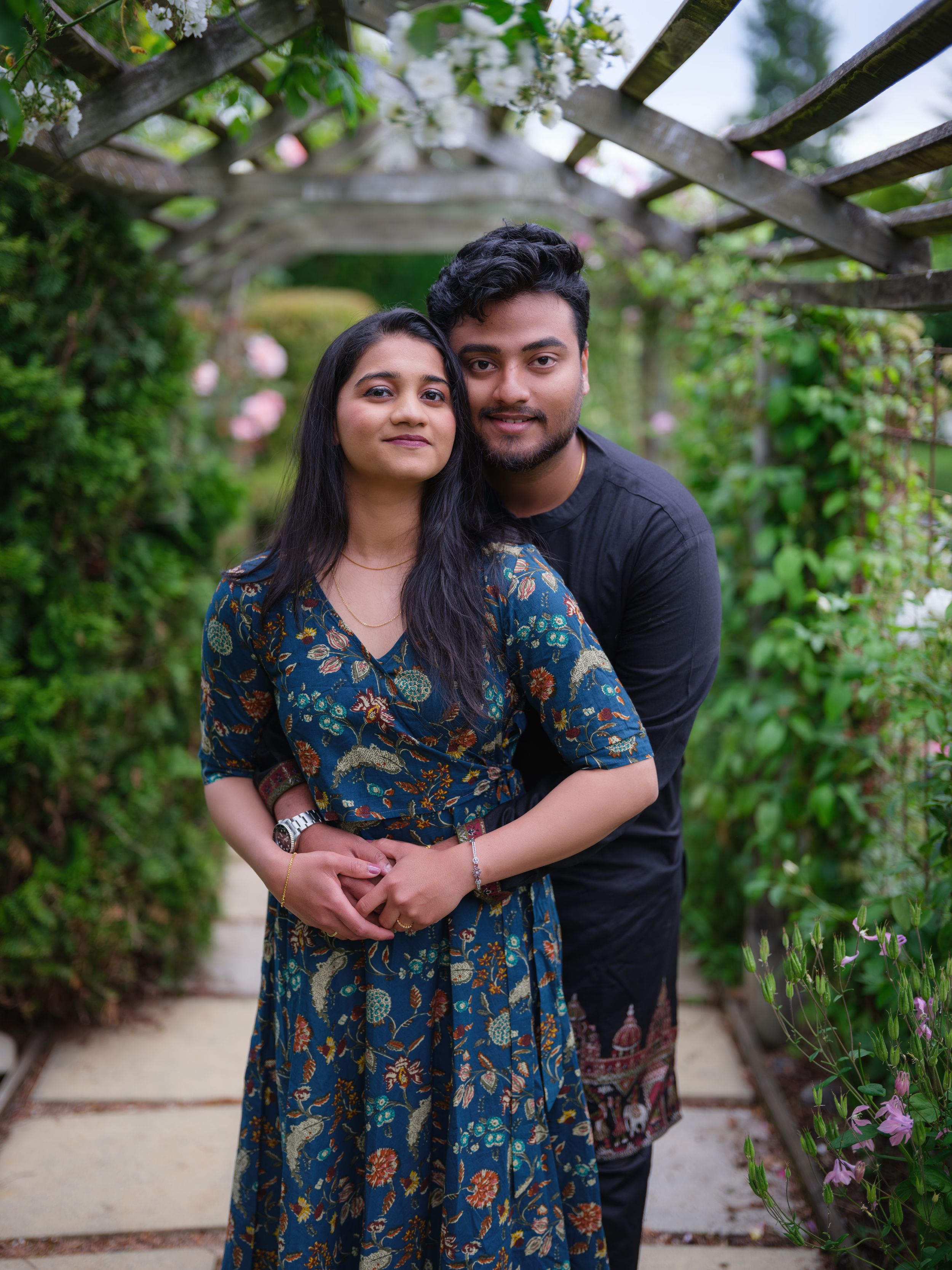 A young couple poses under a floral pergola surrounded by lush greenery, woman in blue floral dress.