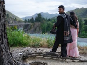 South Asian couple in traditional attire standing by a scenic river with mountains in the background.