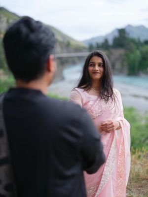 Woman in pink saree smiling at man in black shirt with mountain river bridge backdrop.