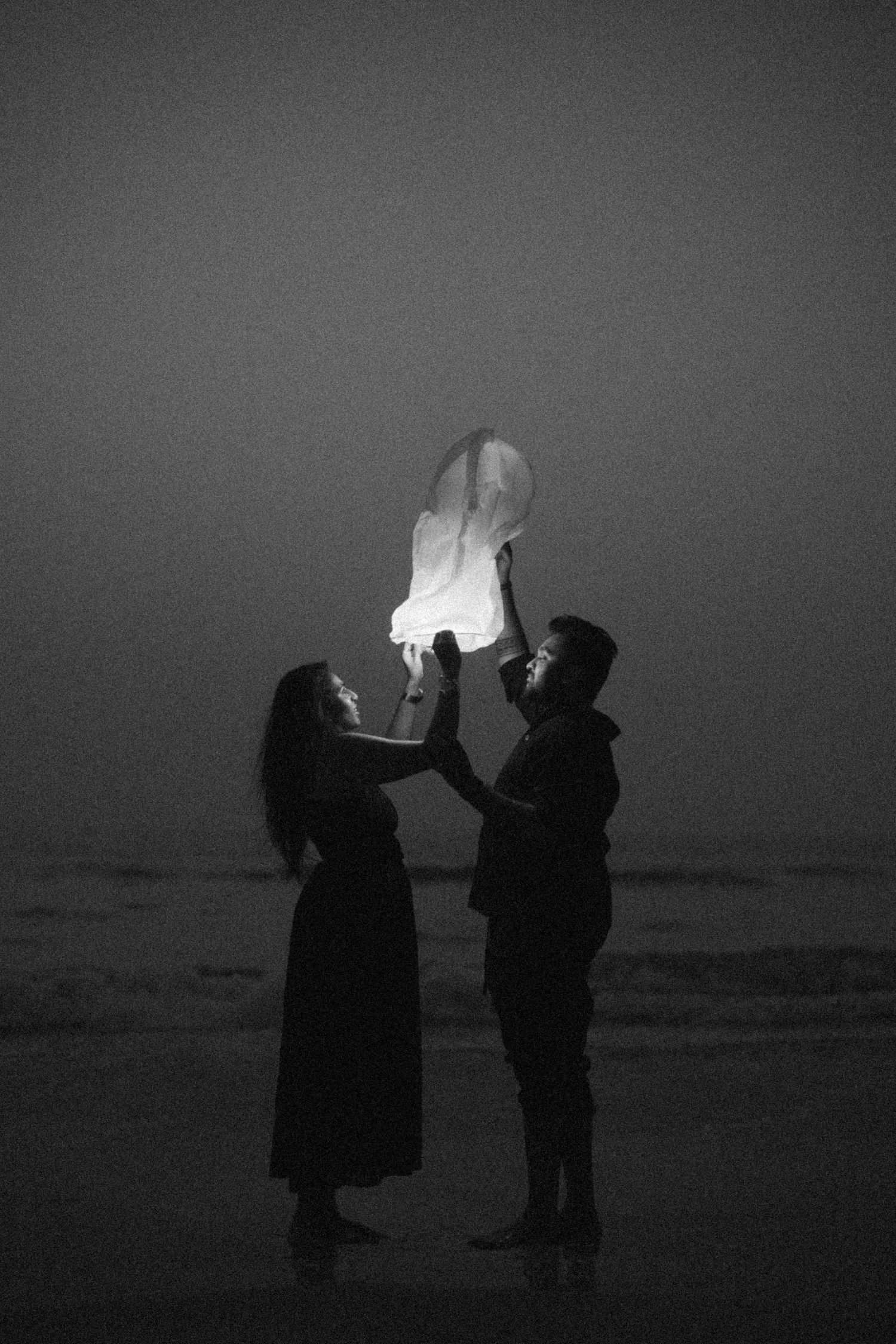 Couple releasing a glowing sky lantern on a beach at dusk, standing by the ocean shoreline.