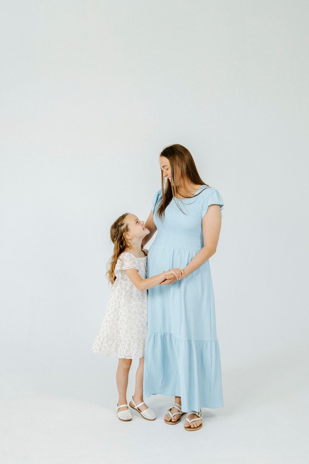 Pregnant woman in blue dress holds hands with young girl in white floral dress against light background.
