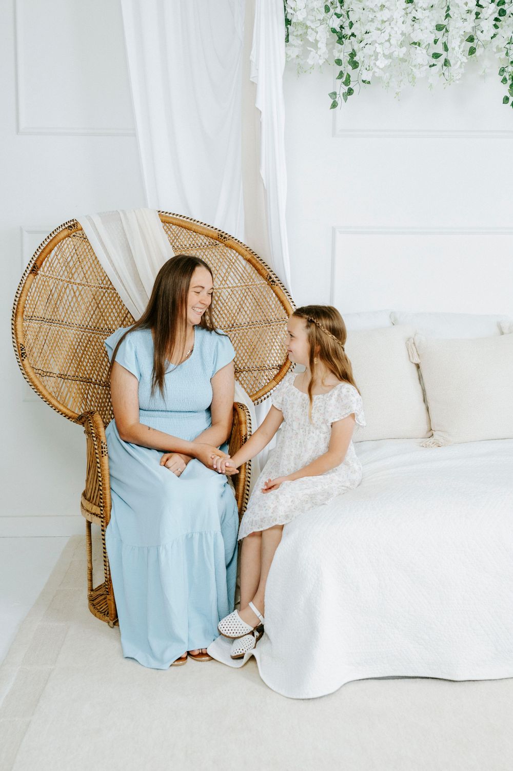 Woman in blue dress sits in wicker chair with young girl in white dress in bright white bedroom.