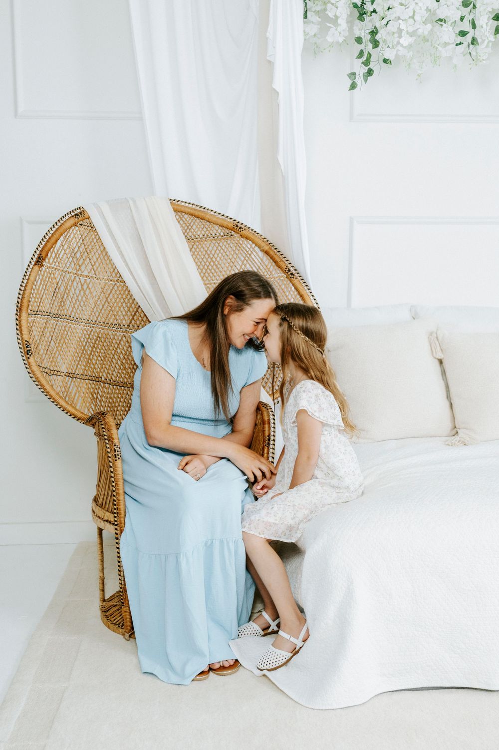 A mother in a blue dress and young girl in white share a tender moment on a bed beside a wicker peacock chair.