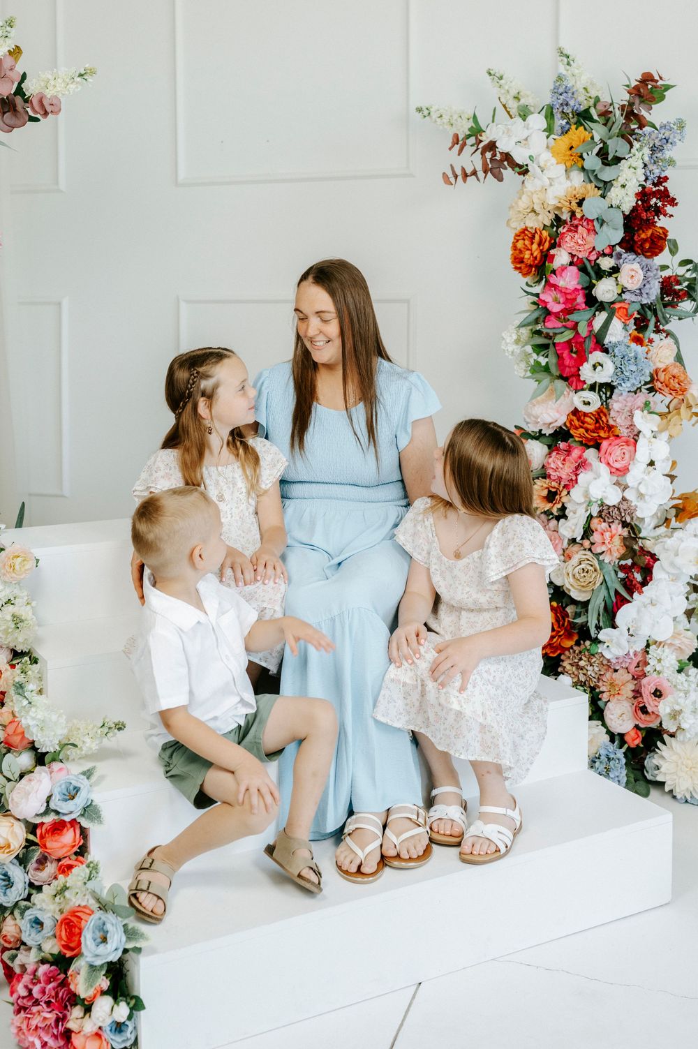 Mother in blue dress smiles with three children surrounded by colorful floral arrangements.