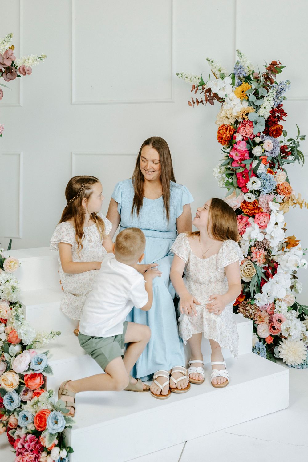 A mother in a light blue dress sits with her three children surrounded by colorful floral arrangements in a bright studio.