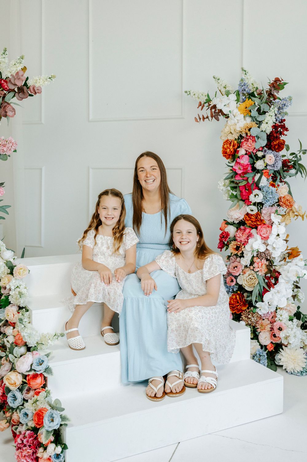 A mother in a light blue dress poses with her two daughters in floral dresses on white steps with colorful flowers.