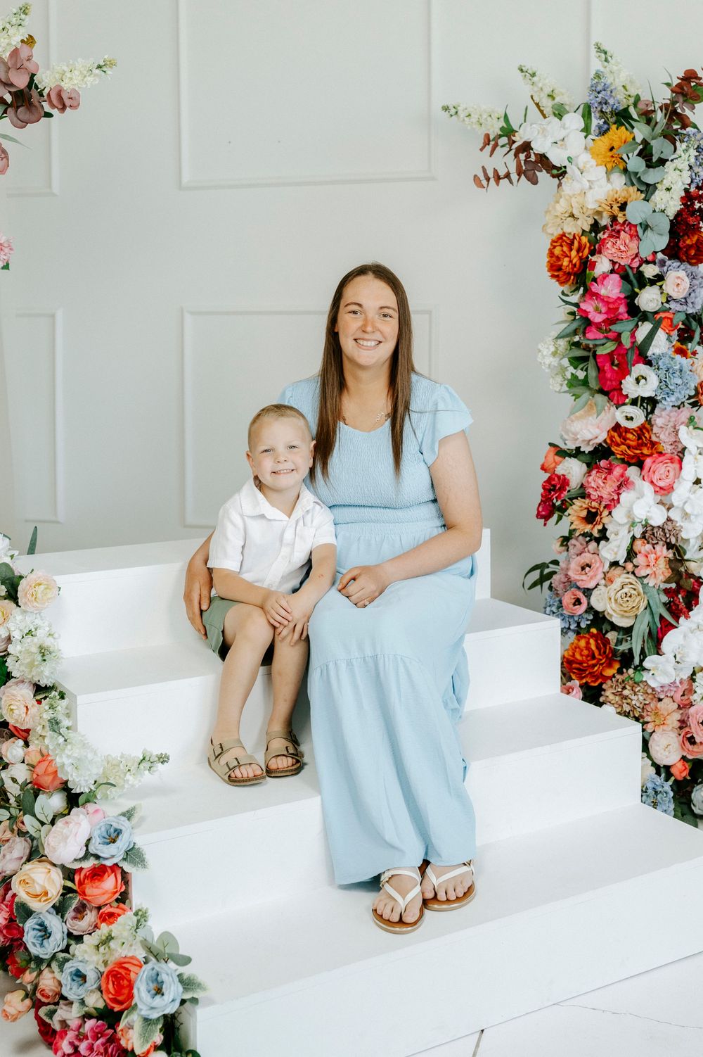 A smiling mother in a light blue dress sits with her young son on white steps surrounded by vibrant floral arrangements.