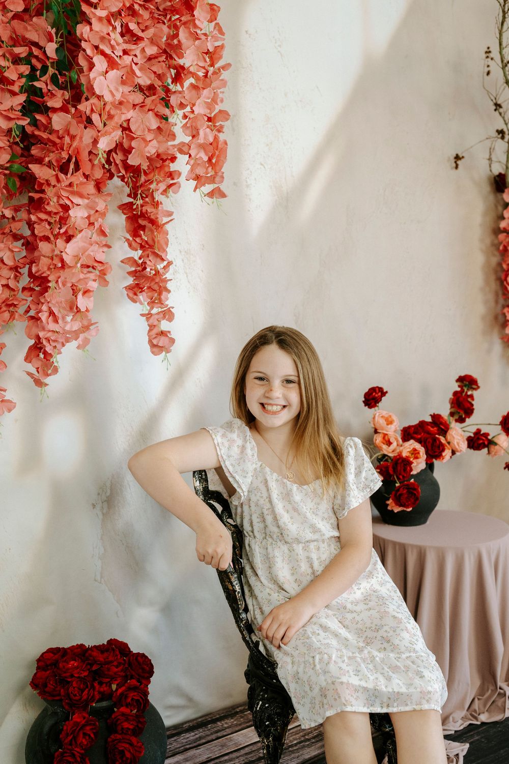 A smiling young girl in a white floral dress sits on a chair surrounded by pink and red flower arrangements.