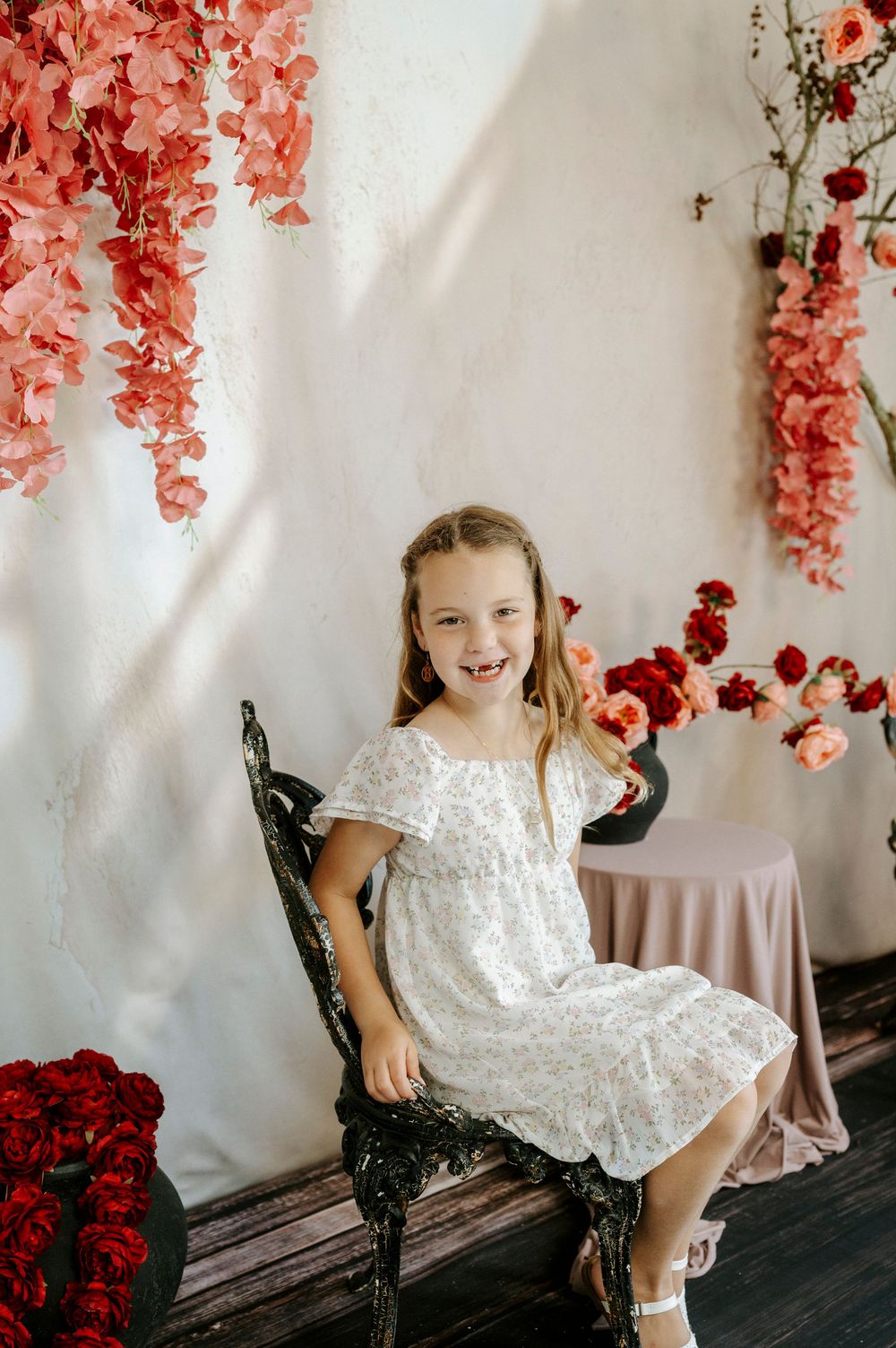 A smiling young girl in a white floral dress sits on a chair surrounded by beautiful pink and red flower arrangements.