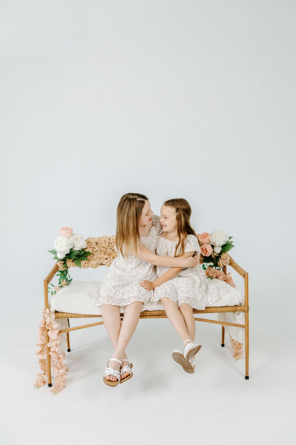 Mother and daughter in matching white dresses sit on a gold bench with pink flowers in a bright studio.