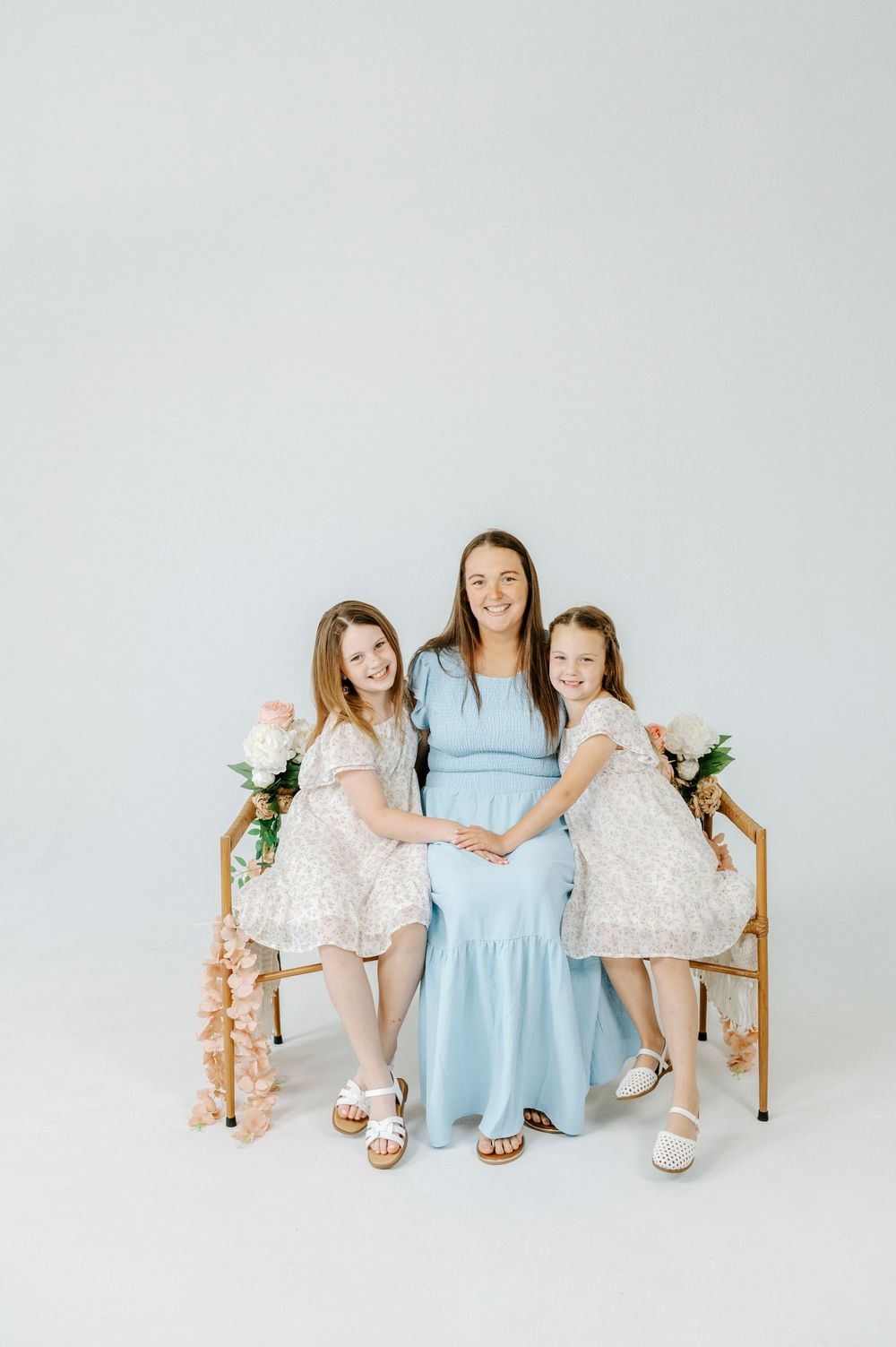 Pregnant mom in blue dress poses with two girls in white dresses on gold bench with florals.
