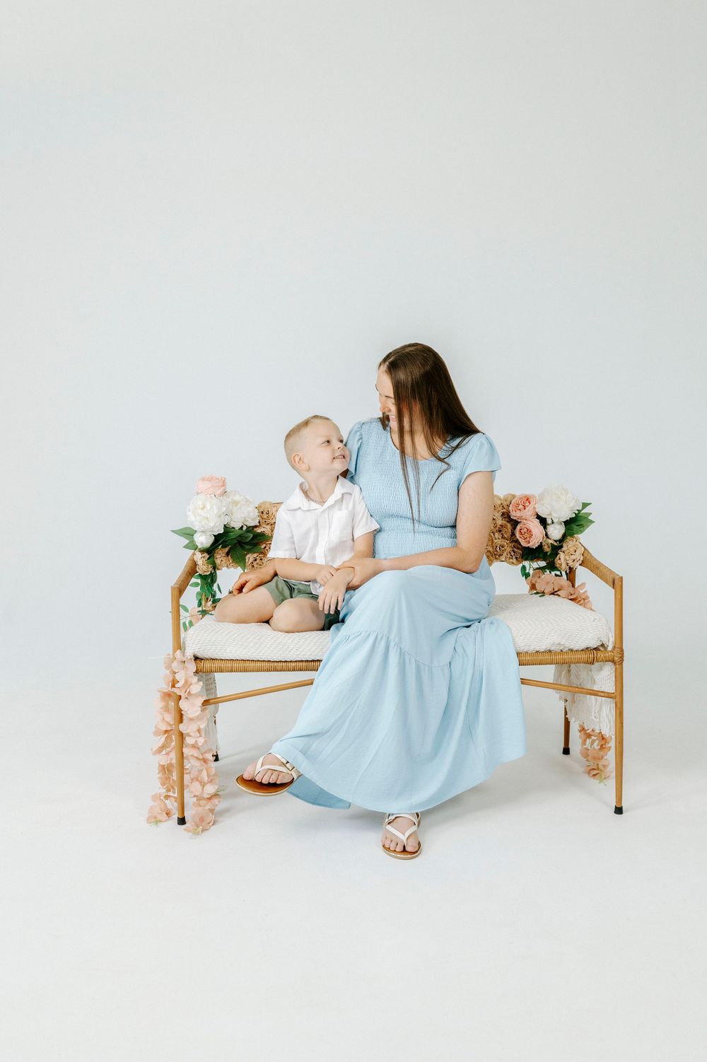 Pregnant woman in blue dress sits on gold bench with toddler boy amid pink and white flowers.