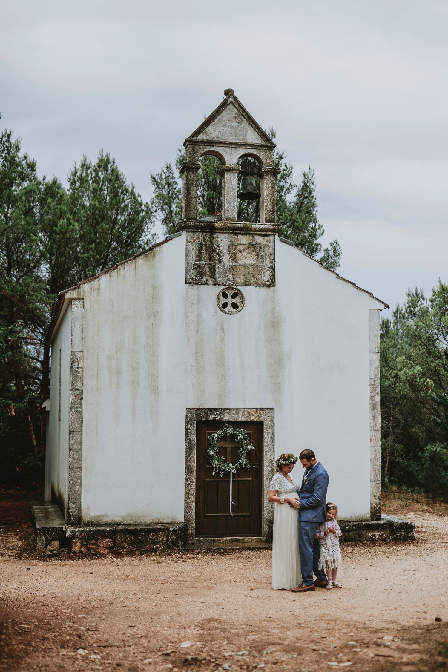 A small white chapel with a rustic bell tower stands against a cloudy sky as a family gathers at its entrance.