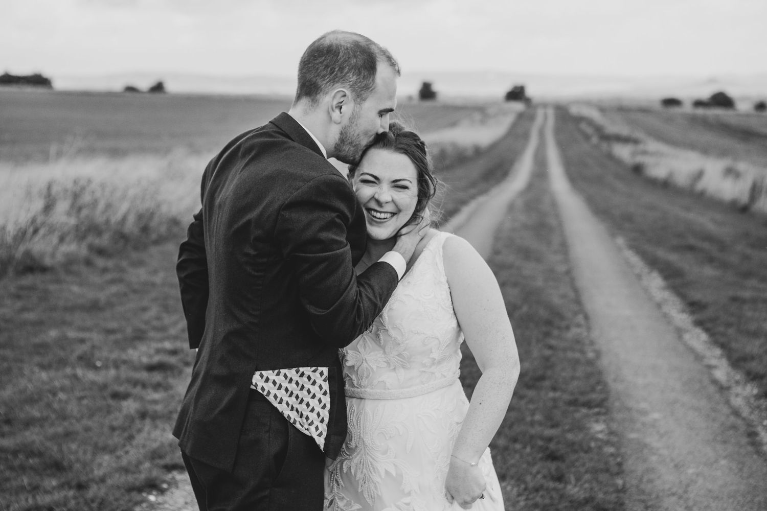 Black and white photo of a smiling bride and groom embracing on a rural dirt path surrounded by open fields.