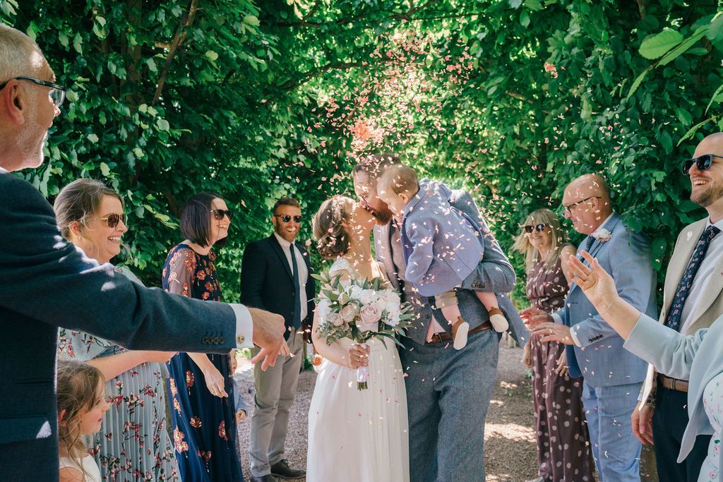 Bride and groom kissing during outdoor wedding confetti celebration, surrounded by guests under lush green tree canopy.