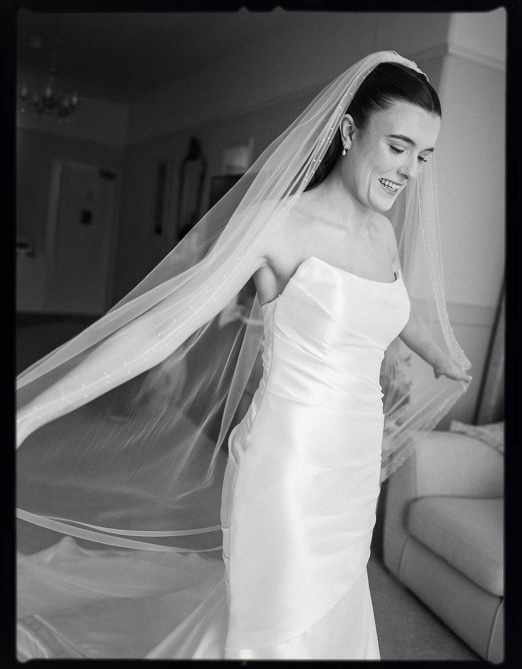 Bride in strapless satin gown and flowing cathedral veil smiles joyfully in black and white wedding portrait.