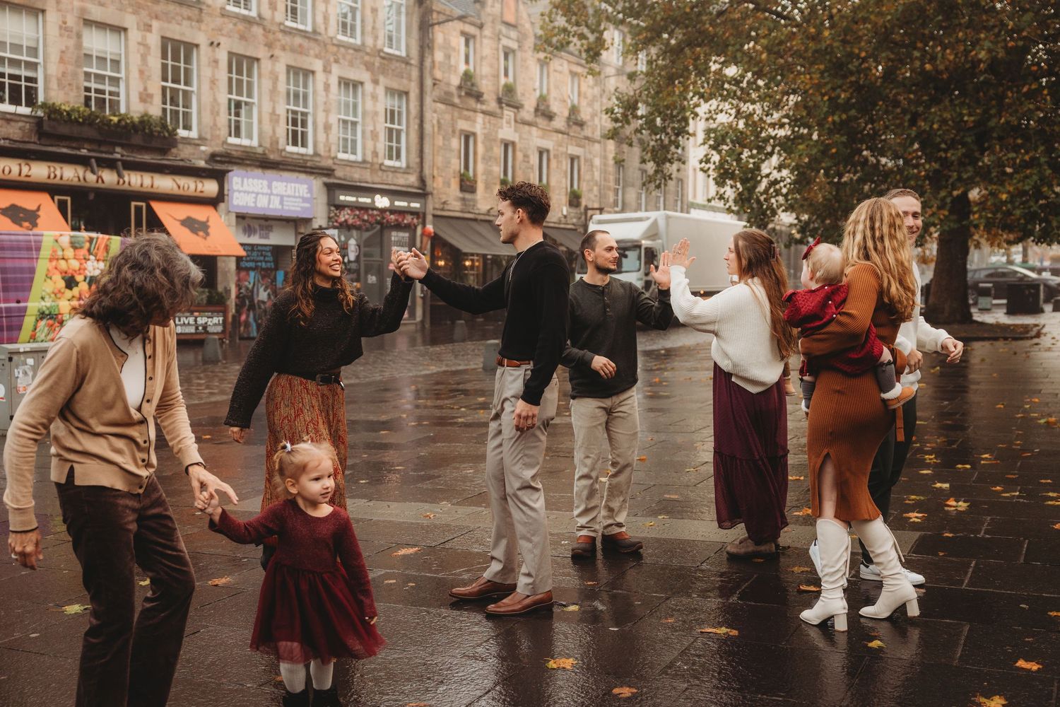 A joyful group of people dancing together on a rainy cobblestone street in Edinburgh city centre.