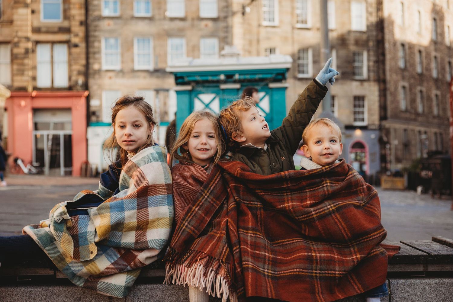 Four children wrapped in plaid tartan blankets pose on a cobblestone street in Edinburgh, Scotland.