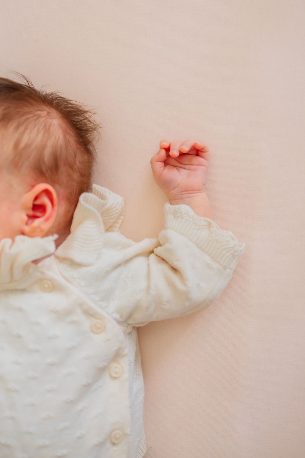 A newborn baby in a white knit outfit lies on a beige surface, with tiny fist raised and ear visible.