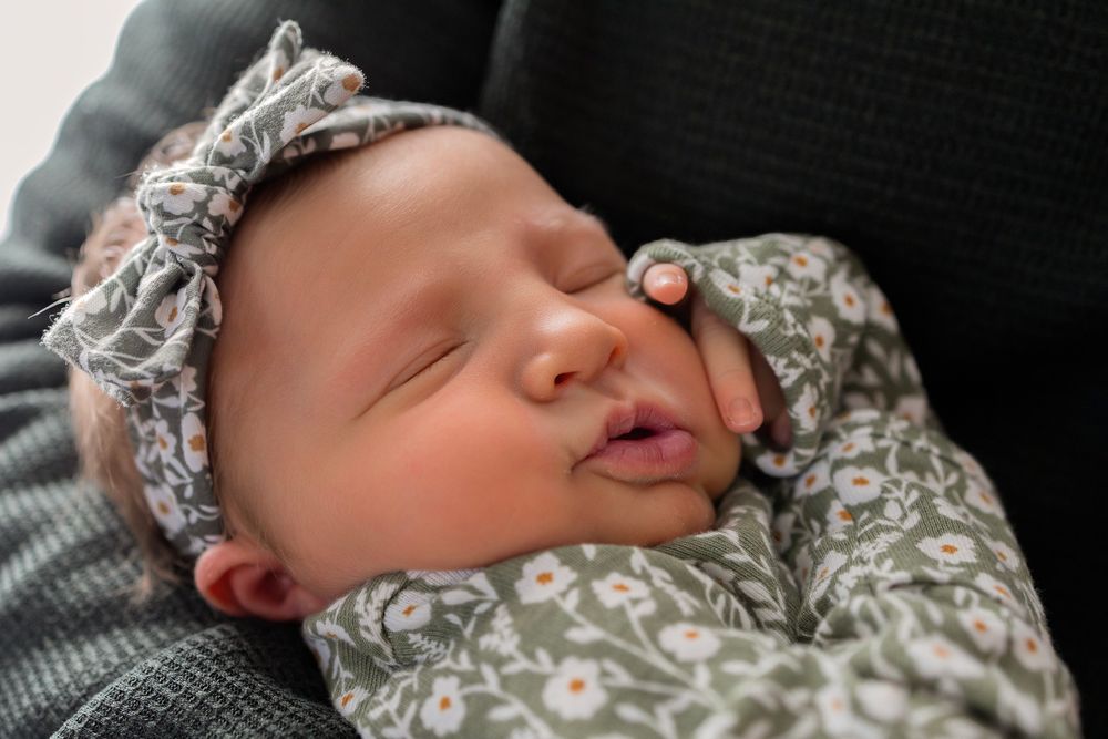 Sleeping newborn baby girl in floral headband and matching green outfit, peacefully resting on a gray surface.