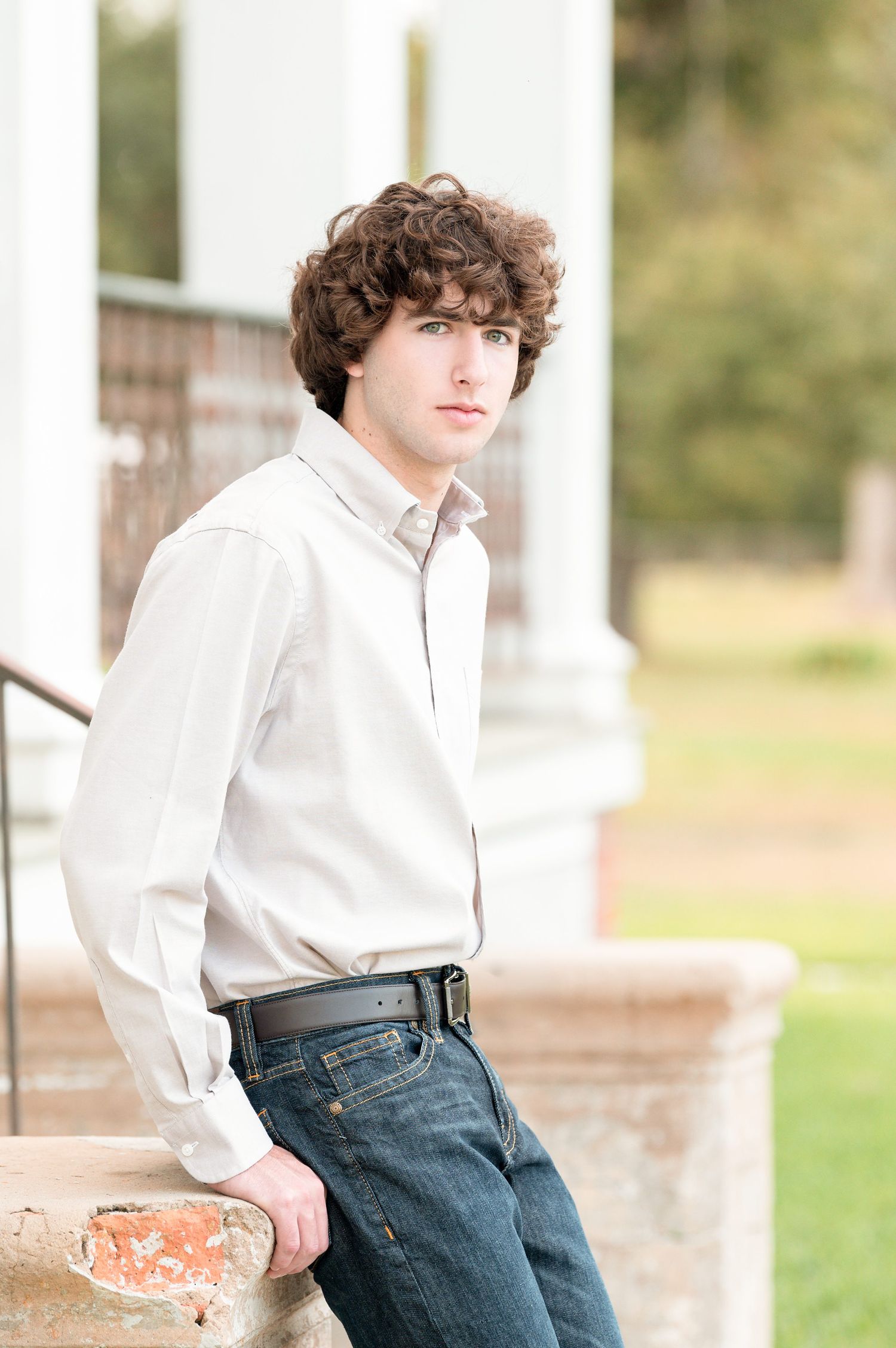 Young man with curly brown hair in white button-up shirt and jeans leaning against brick pillar outdoors.