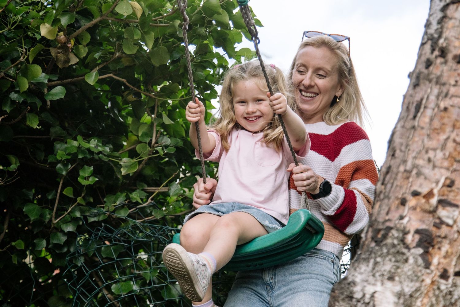 A smiling blonde woman pushes a happy young girl on a green tire swing outdoors among lush green trees.