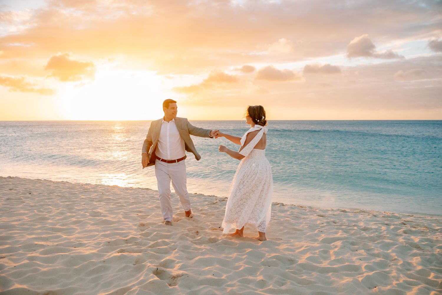 Couple holding hands dancing on a white sandy beach at sunset with turquoise ocean waves in the background.