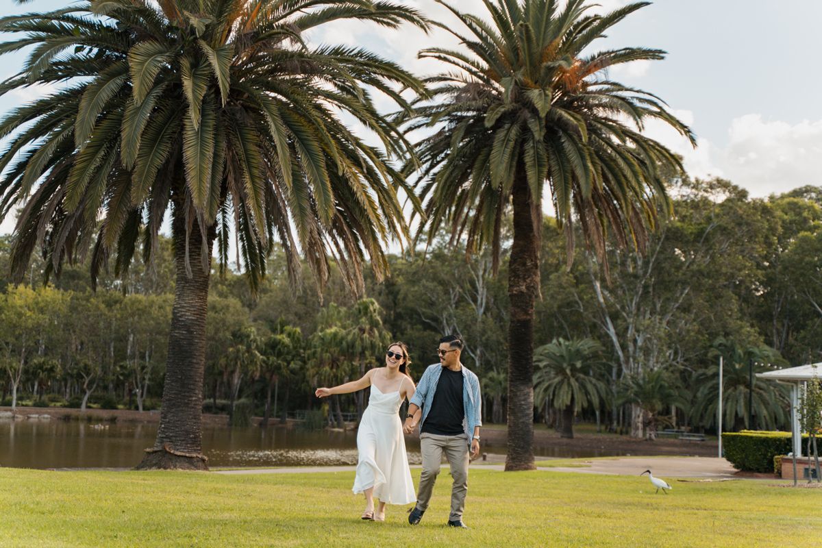 Couple walking hand-in-hand on green lawn with tall palm trees and lake in background during couples photoshoot in western sydney