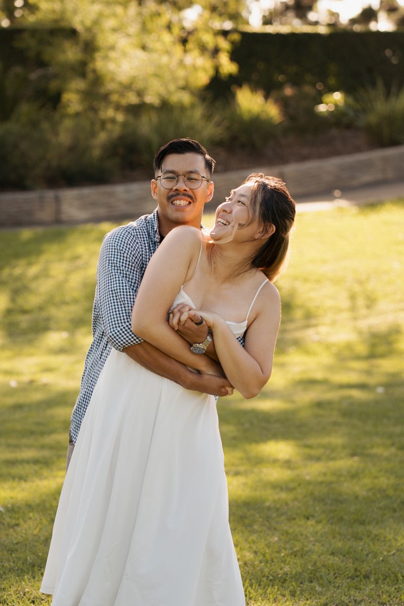 A joyful couple outdoors on green grass, woman in white dress laughing, man in plaid shirt smiling, golden hour light during couples session