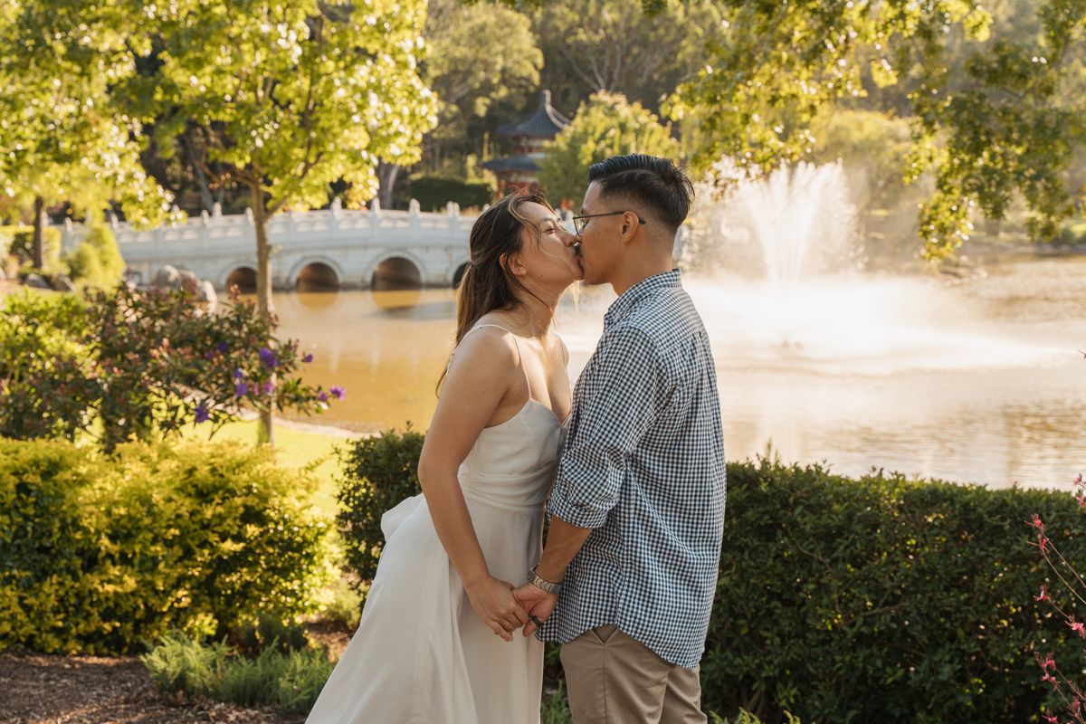 Couple sharing a kiss in a garden with a fountain and pond in the background on a sunny day during an engagement photoshoot with a couple