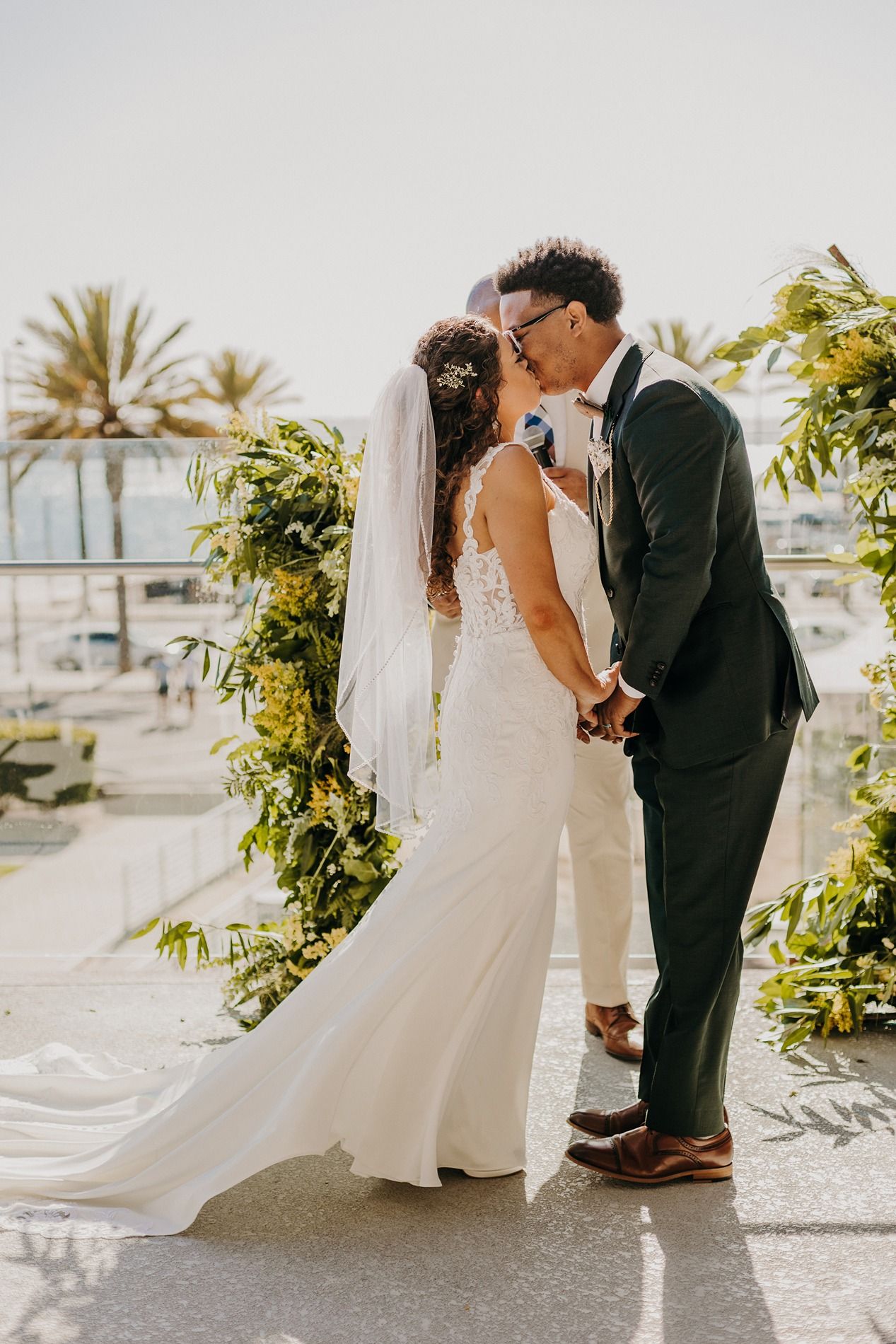 Bride and groom share first kiss at outdoor wedding ceremony with lush greenery arch and palm trees in background at The Lane.