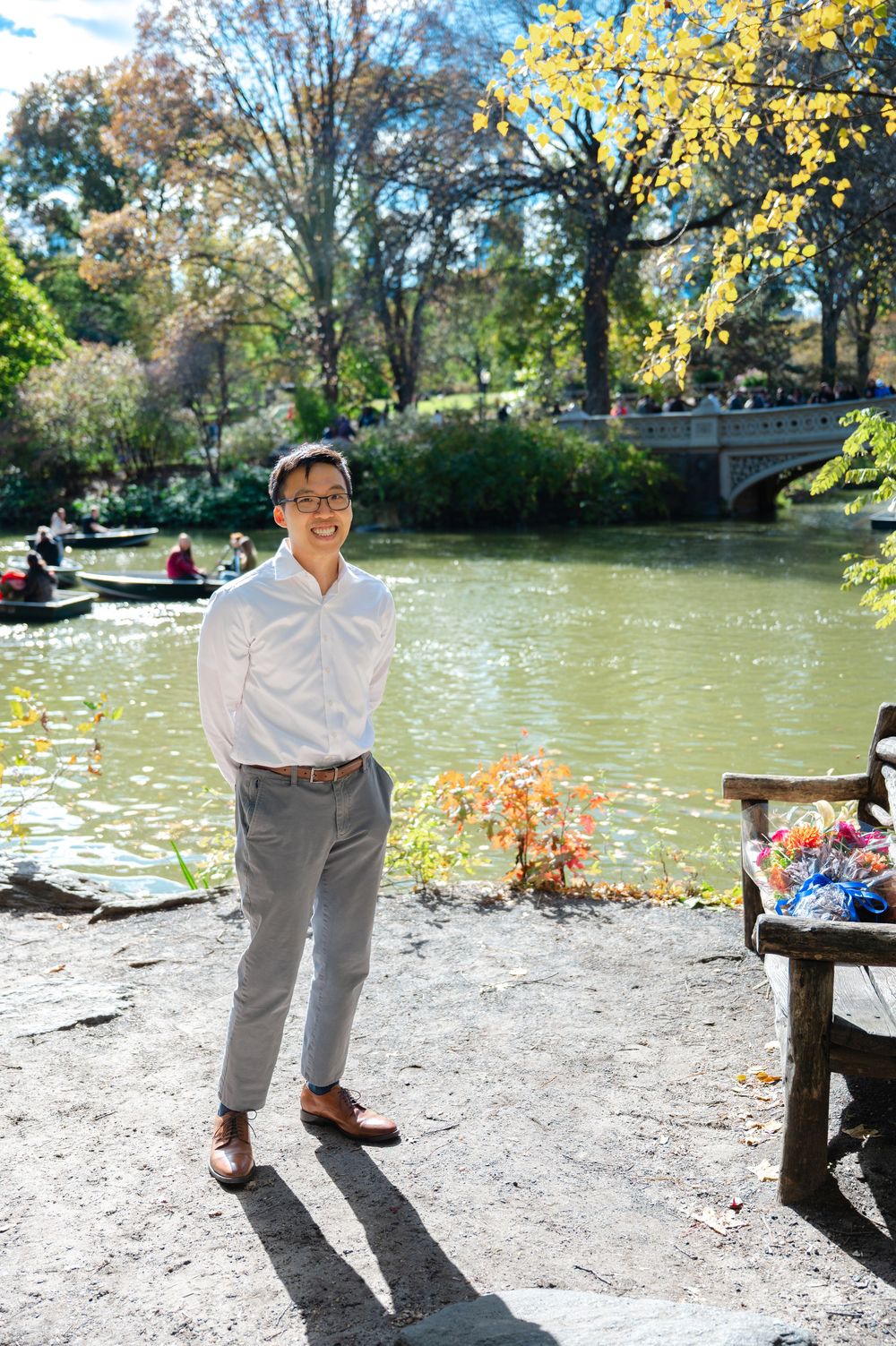 Smiling man in white shirt and gray trousers posing by Central Park lake in fall as he waits to propose to his girlfriend.