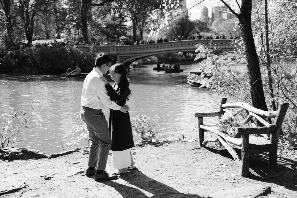 Black and white photo of couple kissing by Central Park lake with Bow Bridge and bench nearby.