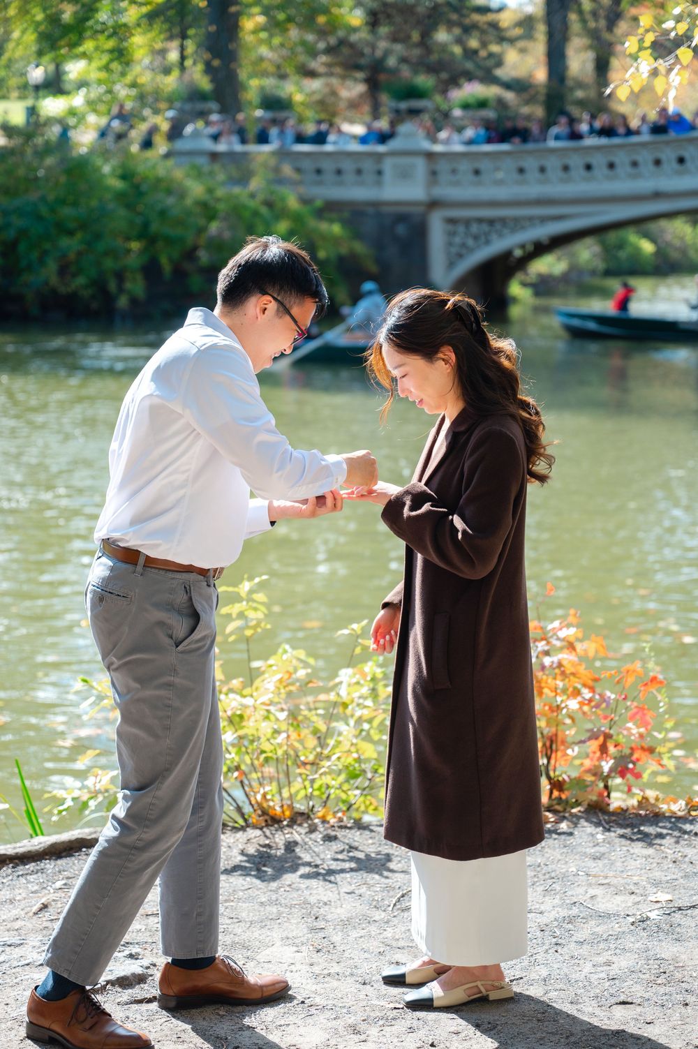 Man placing engagement ring on woman's finger by Central Park lake during autumn proposal.