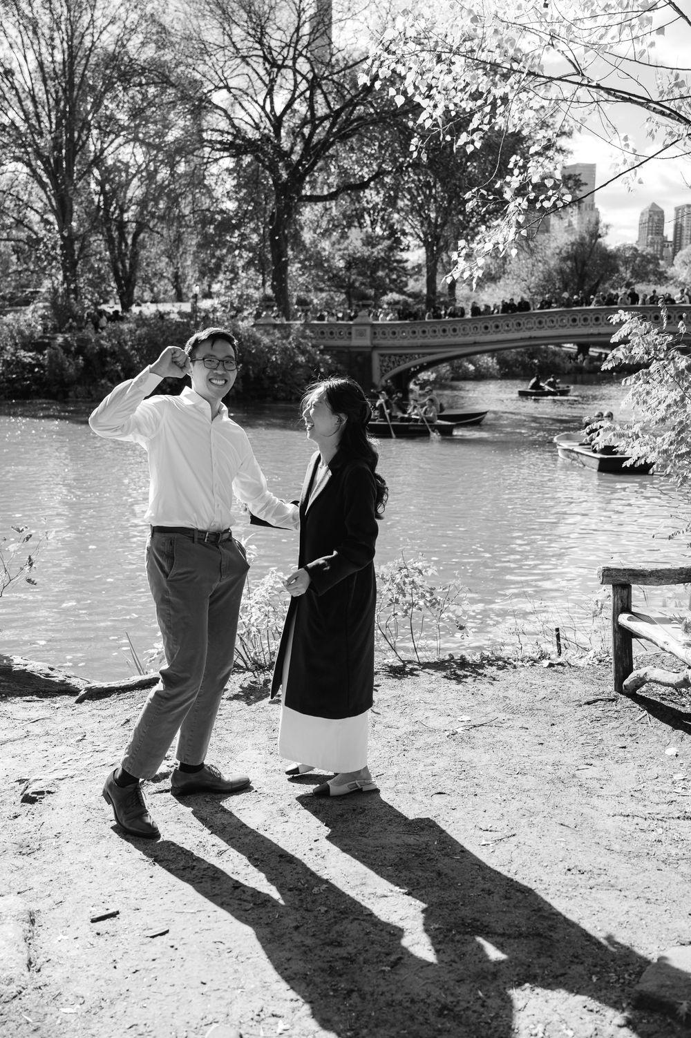 Black and white photo of laughing couple holding hands by Central Park lake with Bow Bridge.