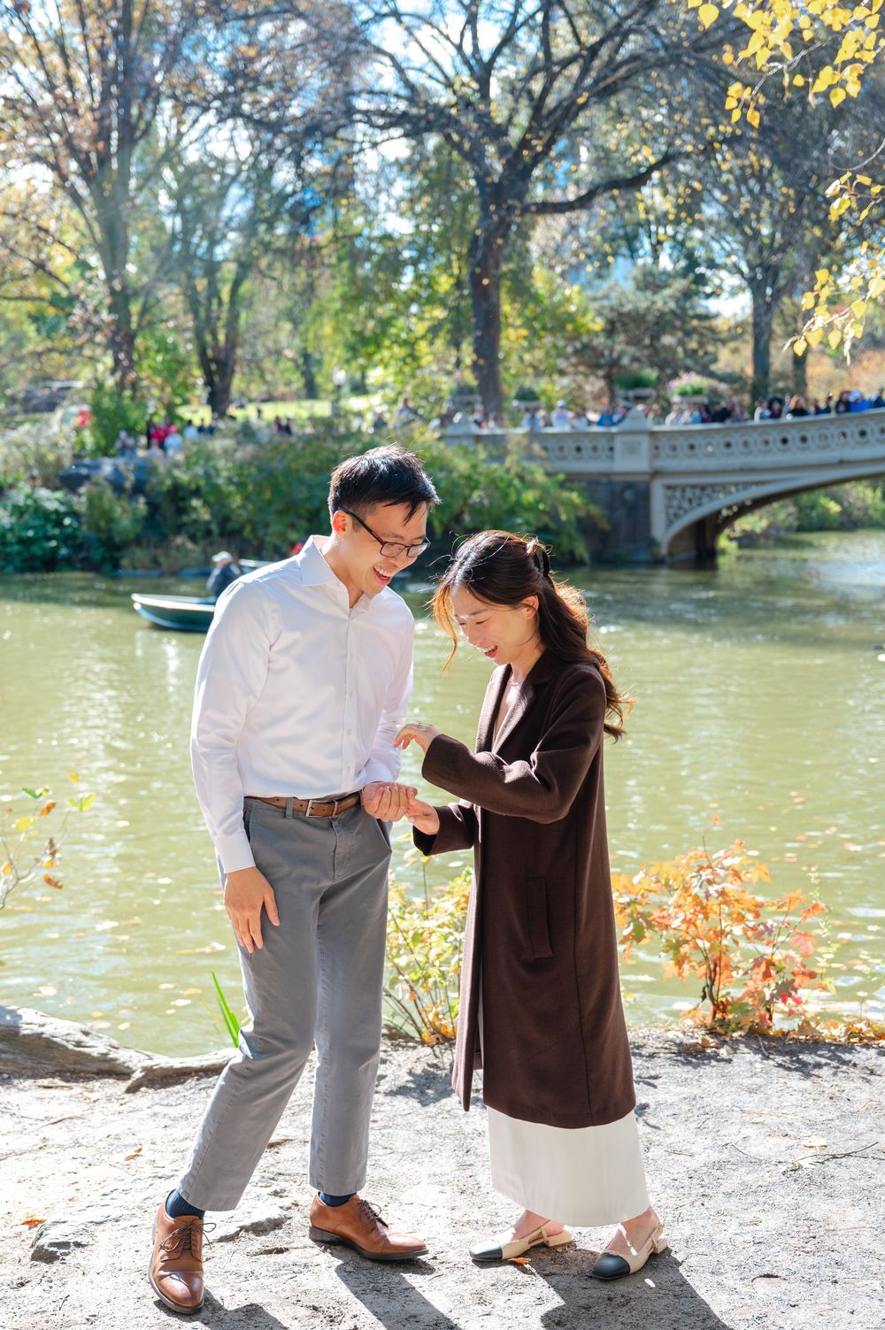 Couple examining engagement ring by Central Park lake with Bow Bridge in background on sunny autumn day.