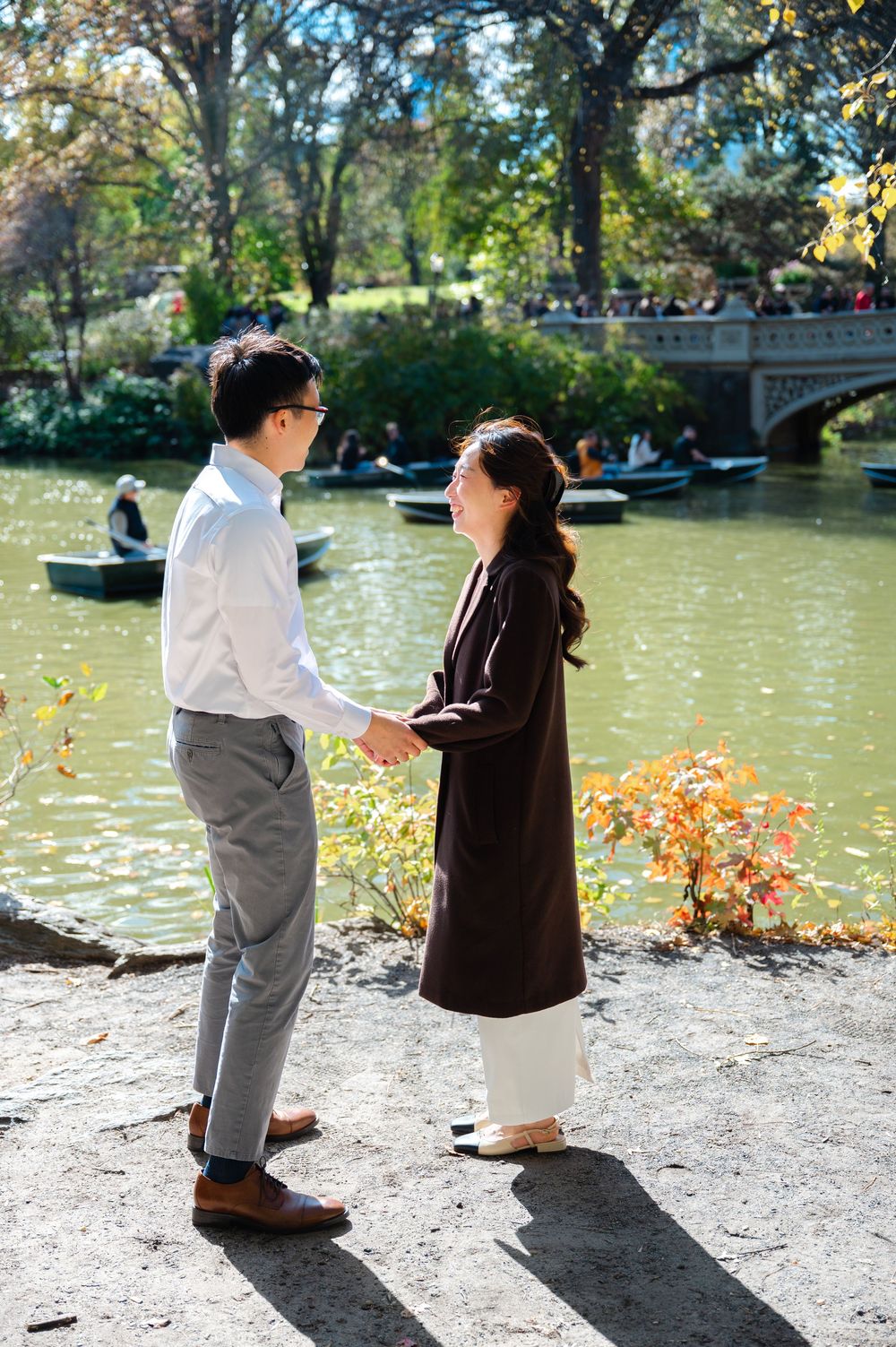 Couple holds hands by Central Park lake with Bow Bridge and rowboats in autumn background.
