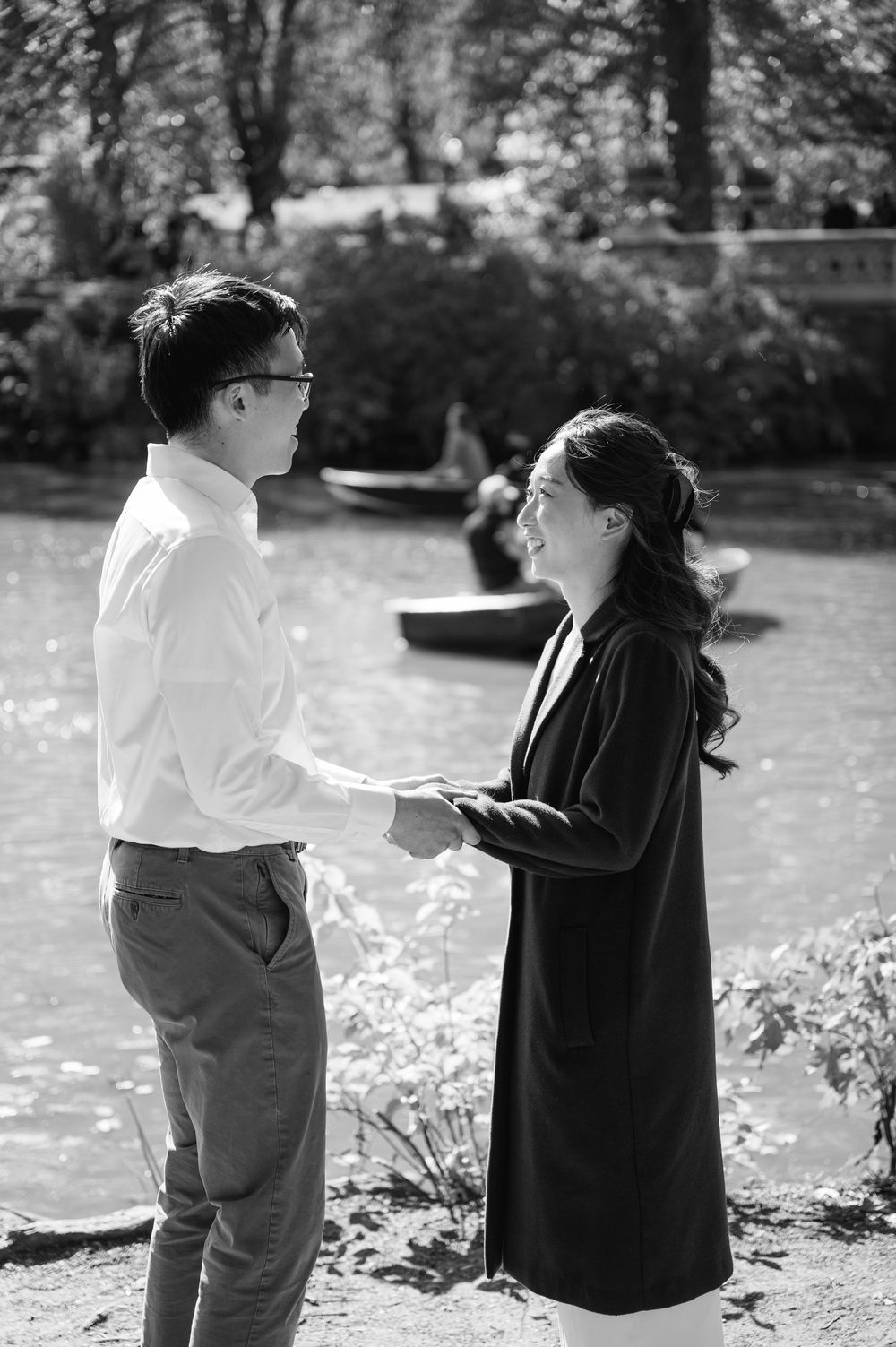 Black and white photo of couple holding hands and exchanging vows by a sunlit lake in Central Park.
