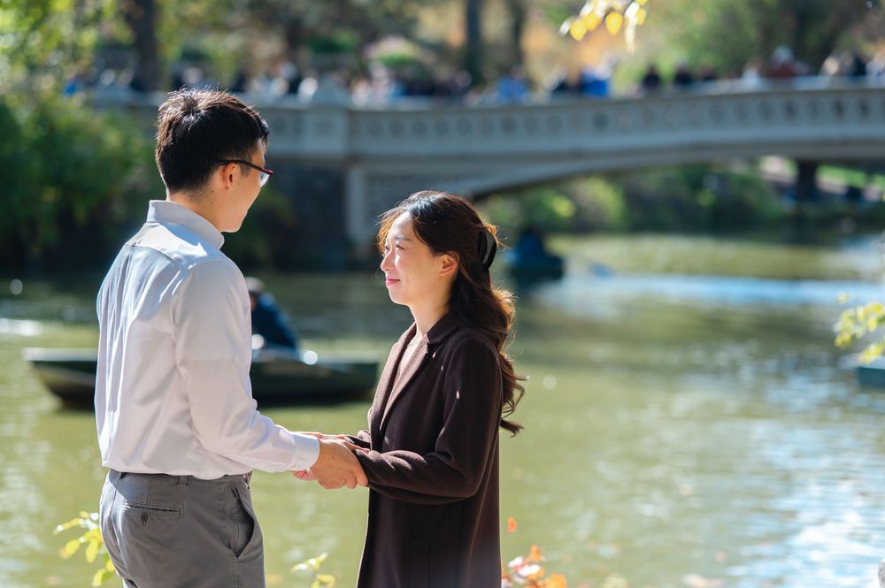 Happy couple holding hands near Central Park lake on a sunny fall day with Bow Bridge behind.