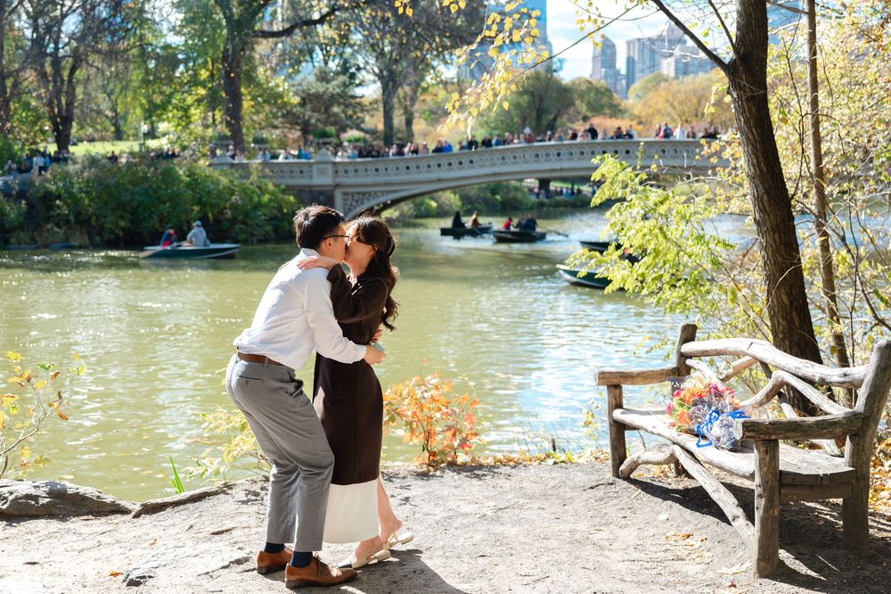 Asian couple sharing a kiss near a scenic Central Park lake with Bow Bridge and rowboats in background.