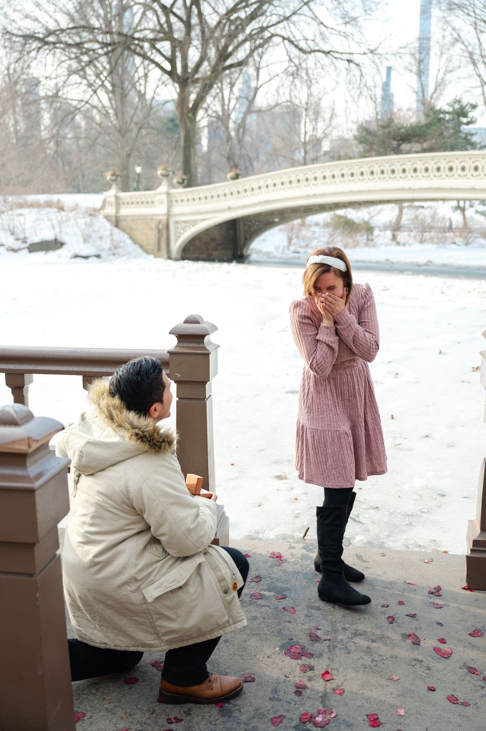Man kneeling on one knee proposing to emotional woman in pink dress near snowy Central Park Bow Bridge.