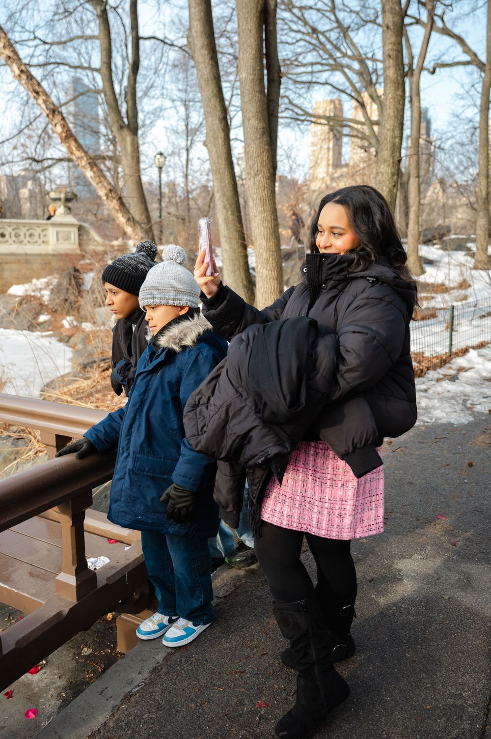 Woman in black puffer jacket holds phone up while two children lean on railing in snowy Central Park.