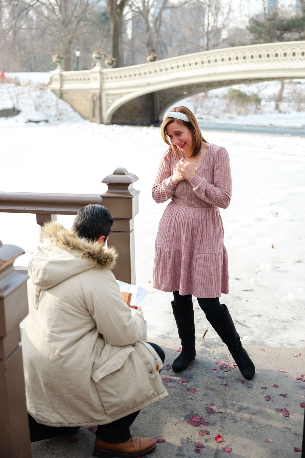 Man proposing to emotional woman in pink dress near snowy Central Park Bow Bridge in winter.