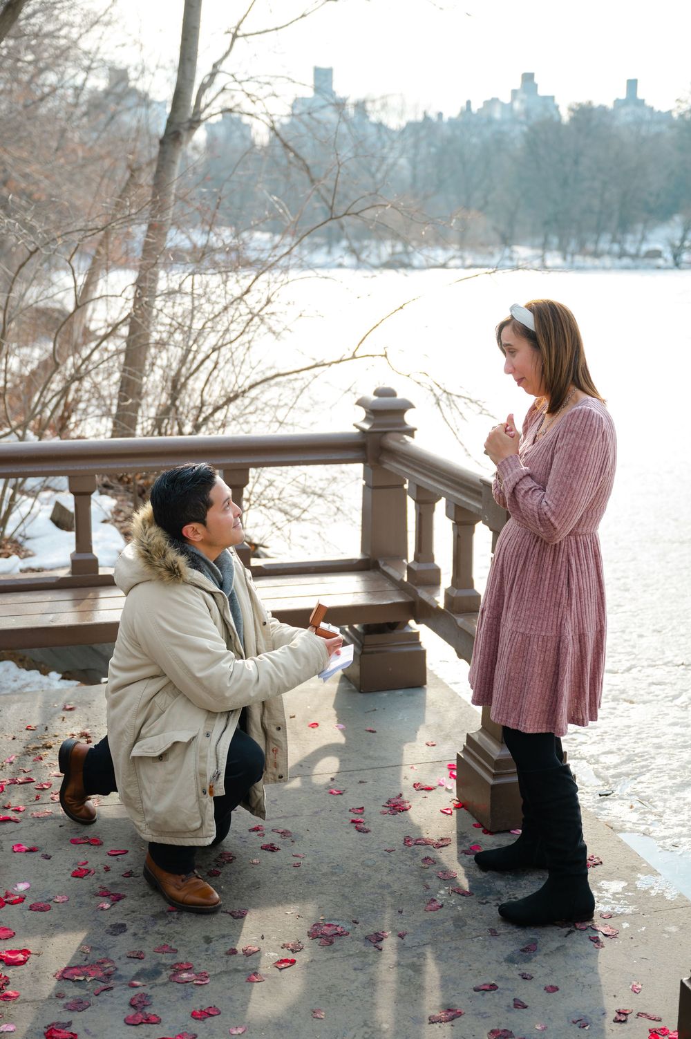 Man proposes to woman on one knee at Central Park in winter, rose petals scattered on ground near snowy lake.