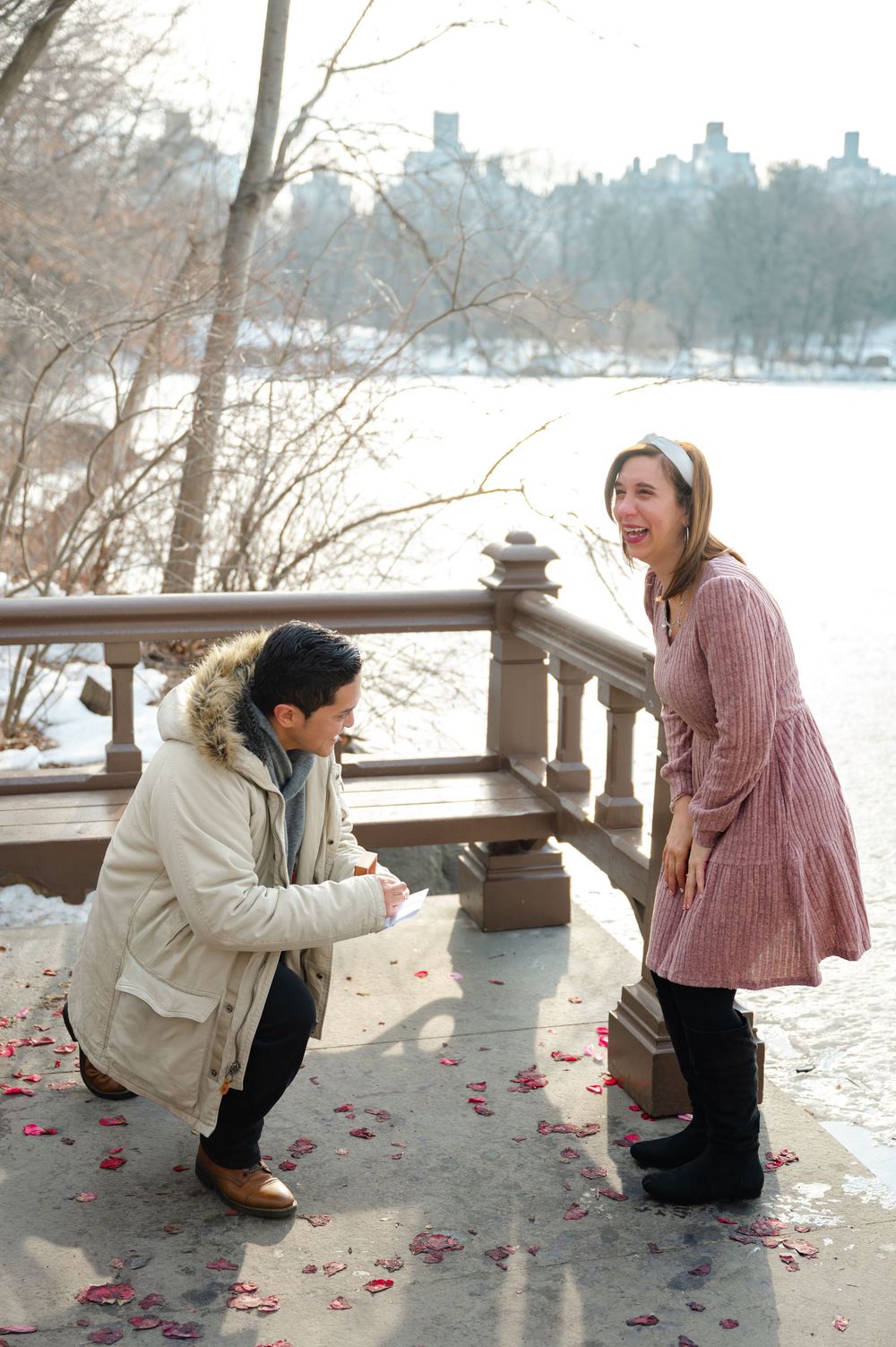 Man proposes to woman on snowy Central Park bridge with rose petals scattered on ground in winter.