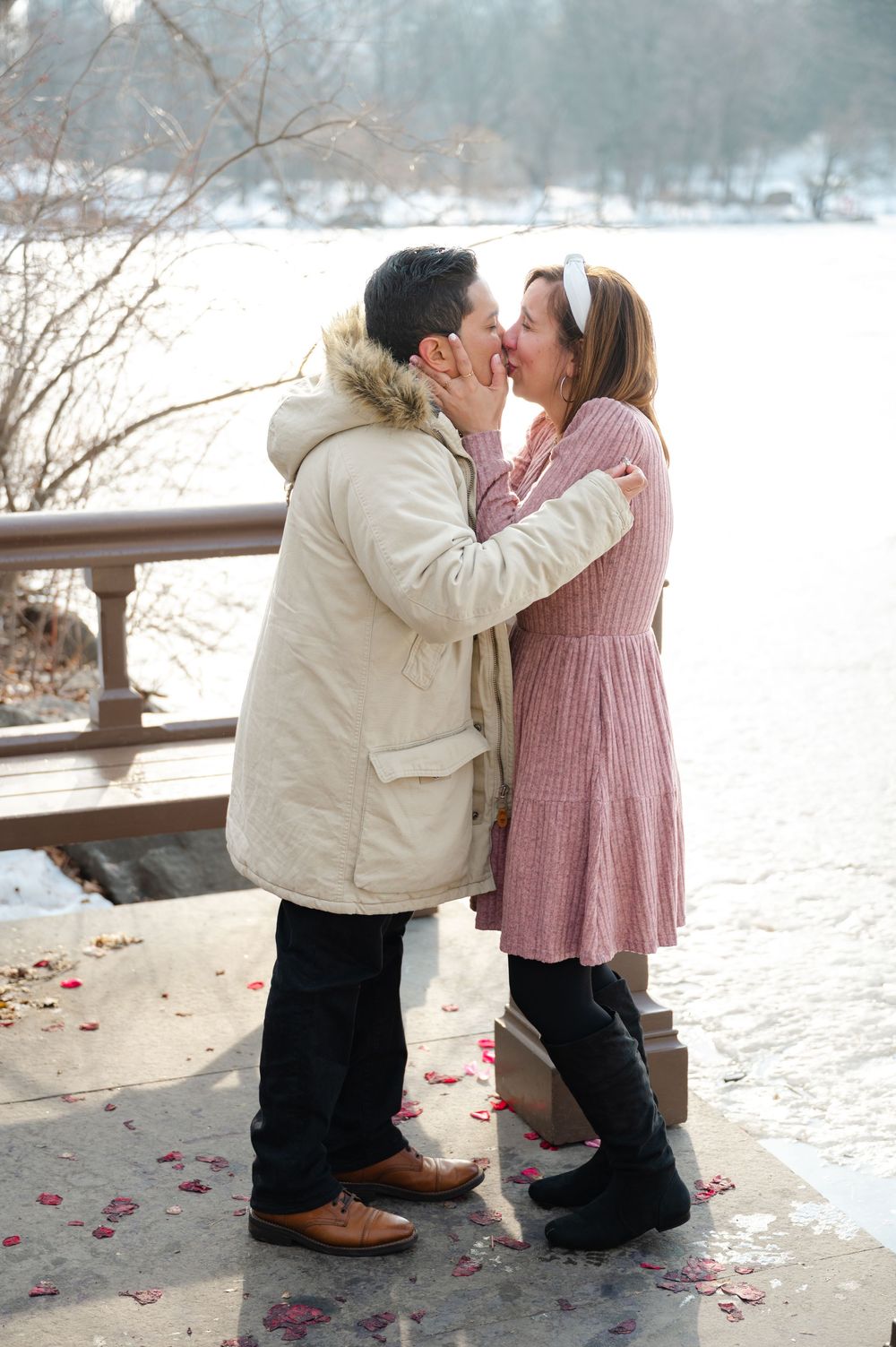 Newly engaged couple sharing first kiss after proposal at snowy Central Park Bow Bridge in winter.