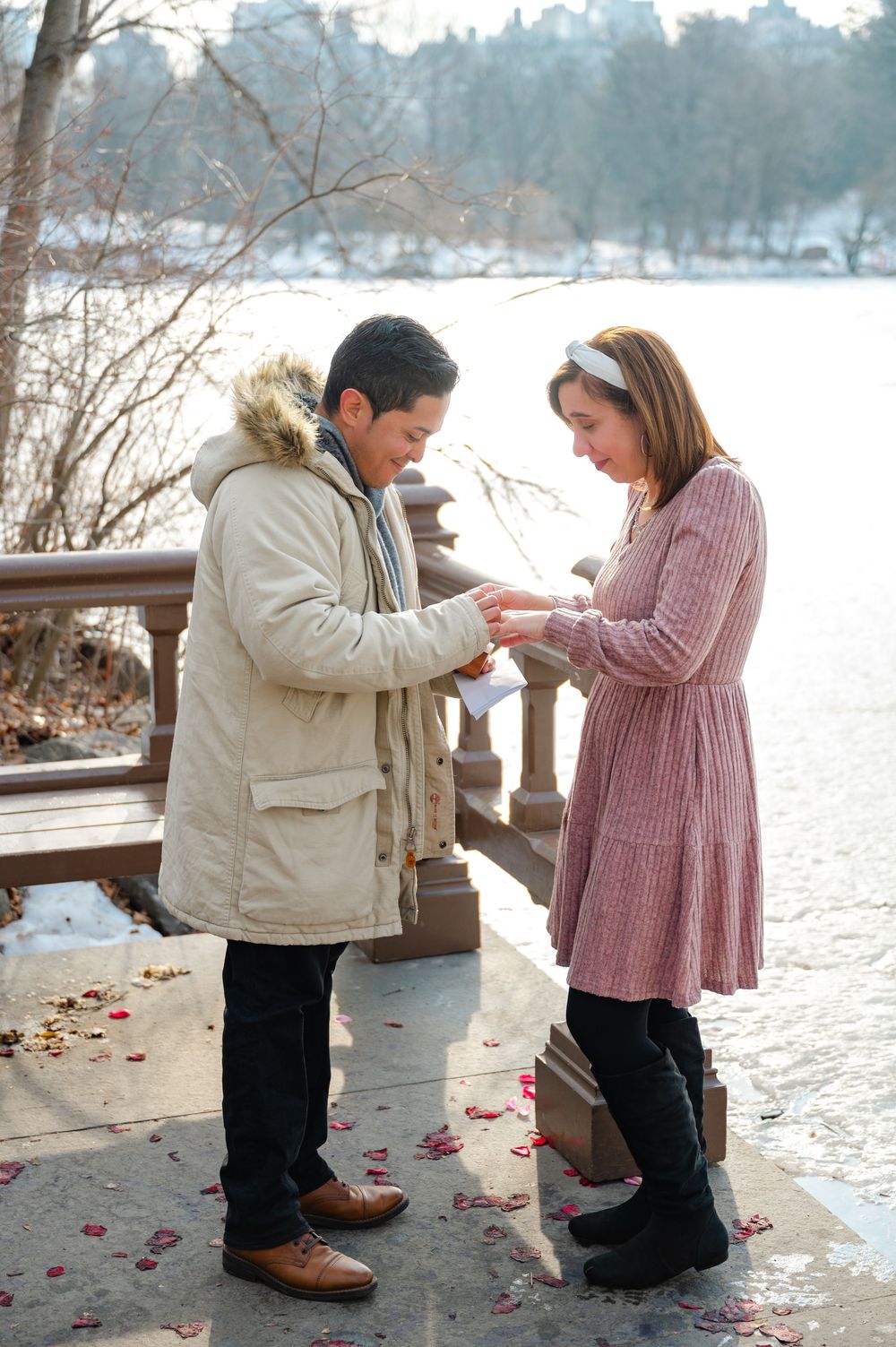 Couple exchanges engagement ring on a snowy Central Park bridge surrounded by scattered rose petals.